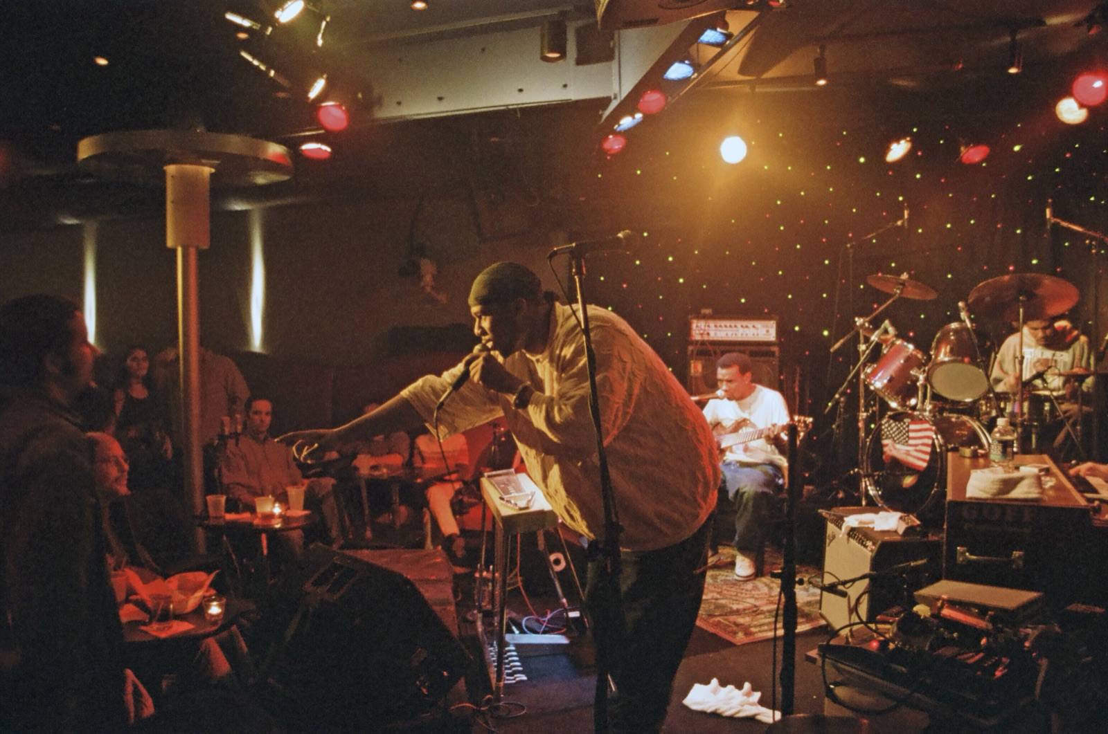 A singer leans out over the club floor with a microphone toward seated fans while the band plays under red and white stage lights.